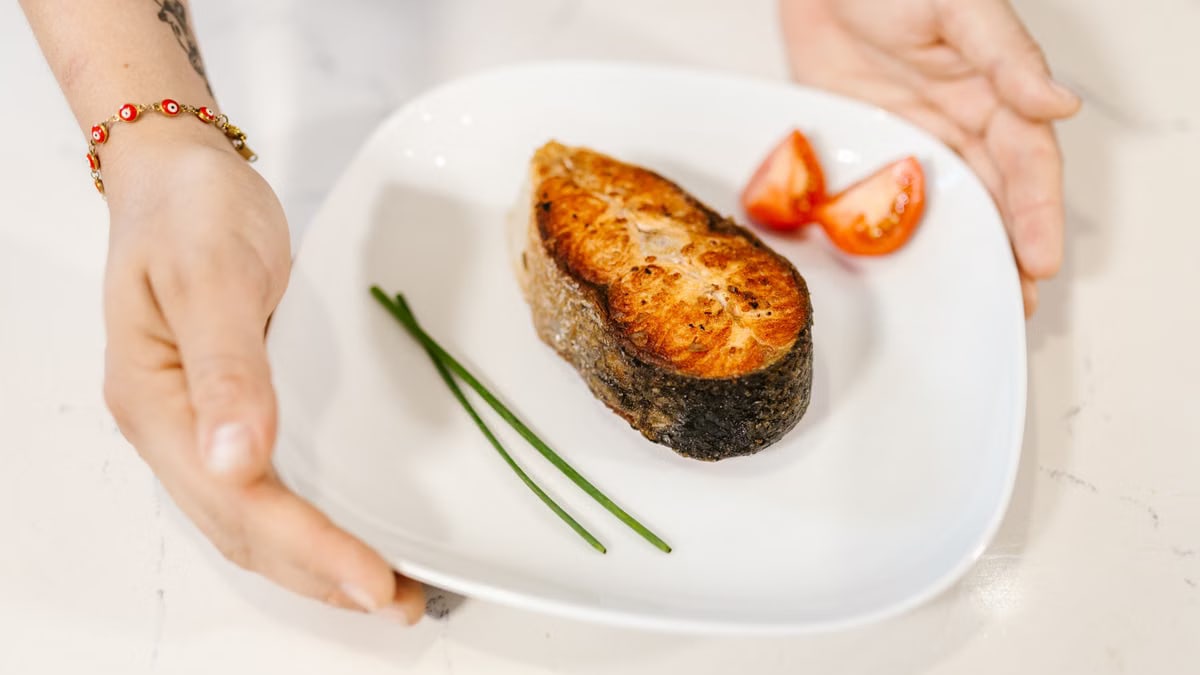 Close-up of grilled salmon served with tomatoes and chives on a plate, held by tattooed hands.